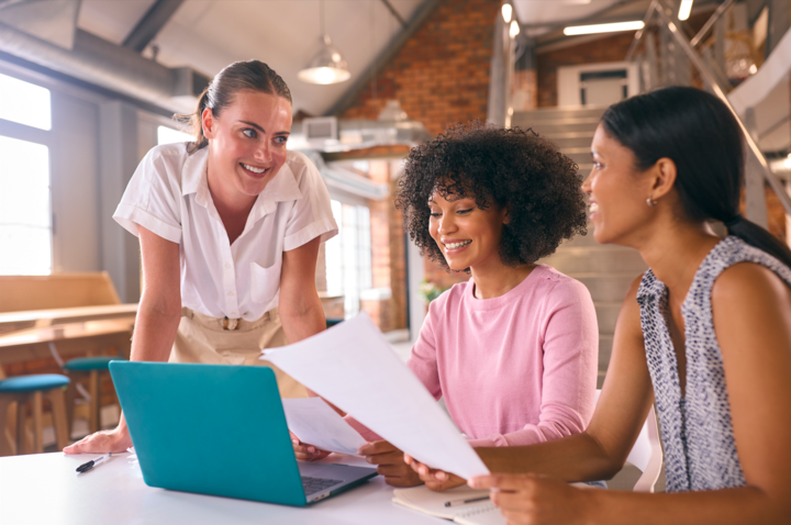 Three Young Businesswomen Meeting In Modern Office Researching Best Payroll Software Sitting Around Table Working On Laptop Together